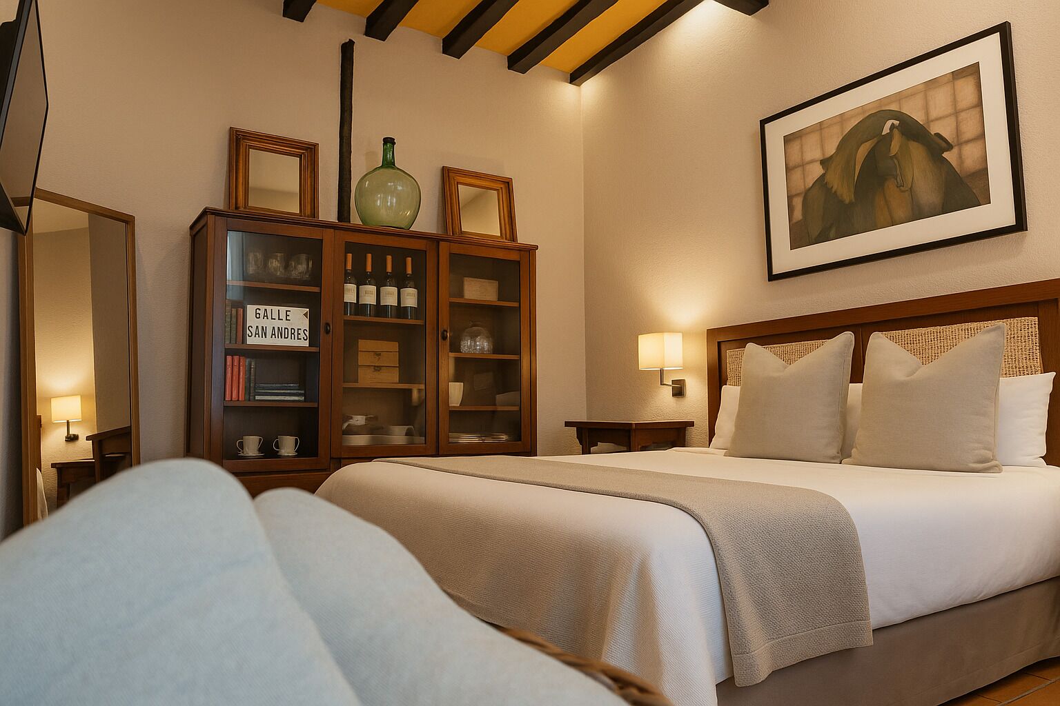 Una habitación de hotel con una cama, un aparador de madera con botellas de vino, un cuadro en la pared y una mesita de noche con una lámpara.