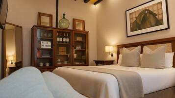 Una habitación de hotel con una cama, un aparador de madera con botellas de vino, un cuadro en la pared y una mesita de noche con una lámpara.