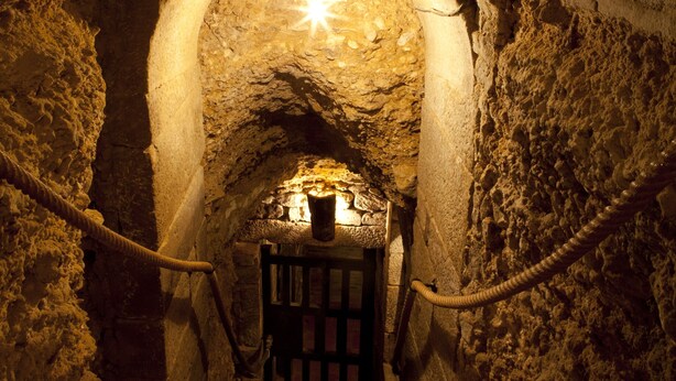A stone tunnel with a wooden door at the end, illuminated by a single hanging light fixture.