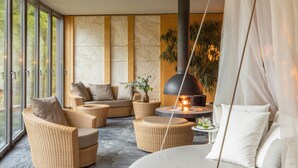 A modern living room with wicker furniture, a fireplace, and large windows offering a view of the outdoors. - Spirit Hotel Thermal Spa (Sarvar)