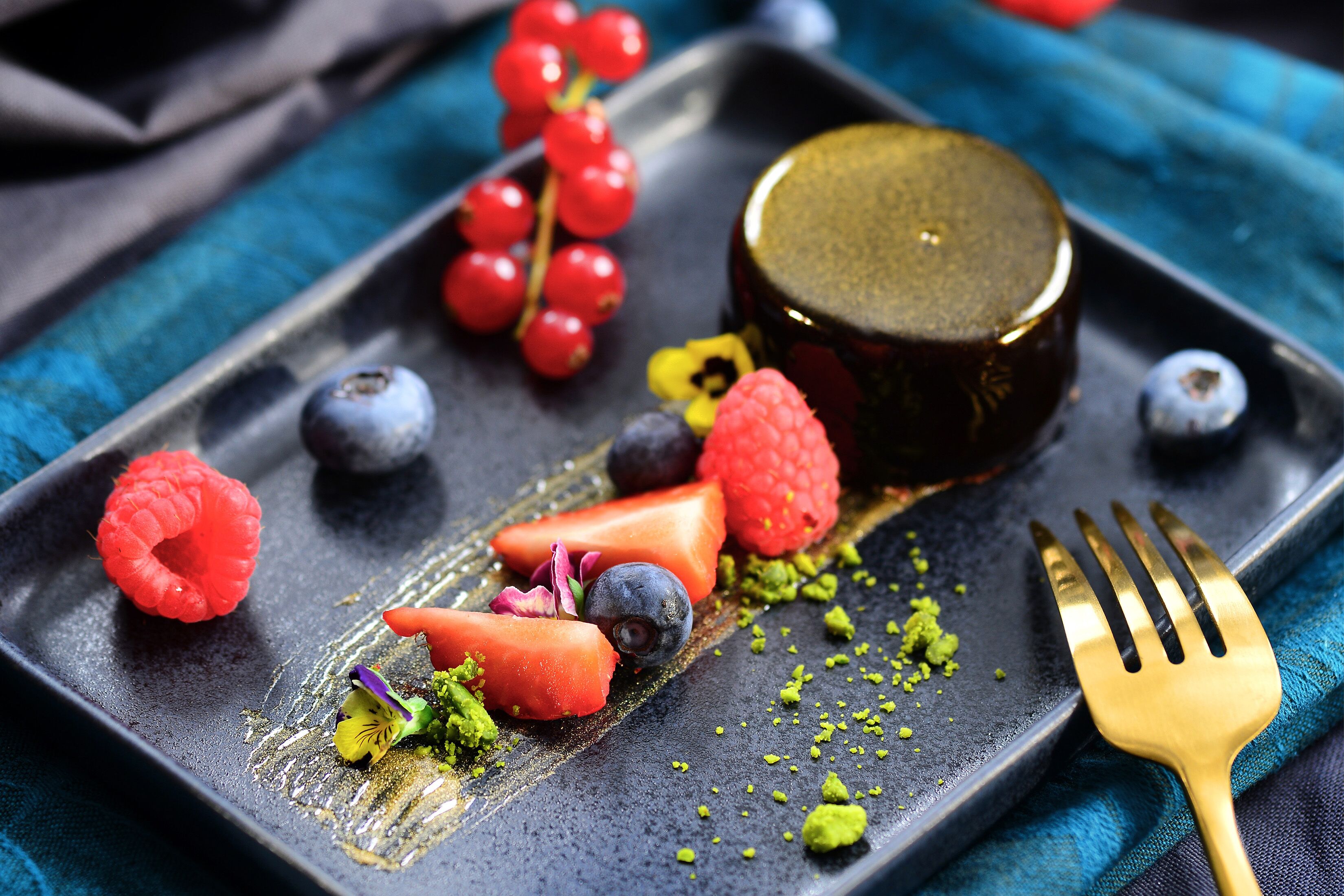 A dessert plate with various fruits and a gold-colored item, possibly a chocolate or caramel, on a dark plate with a fork on the side.