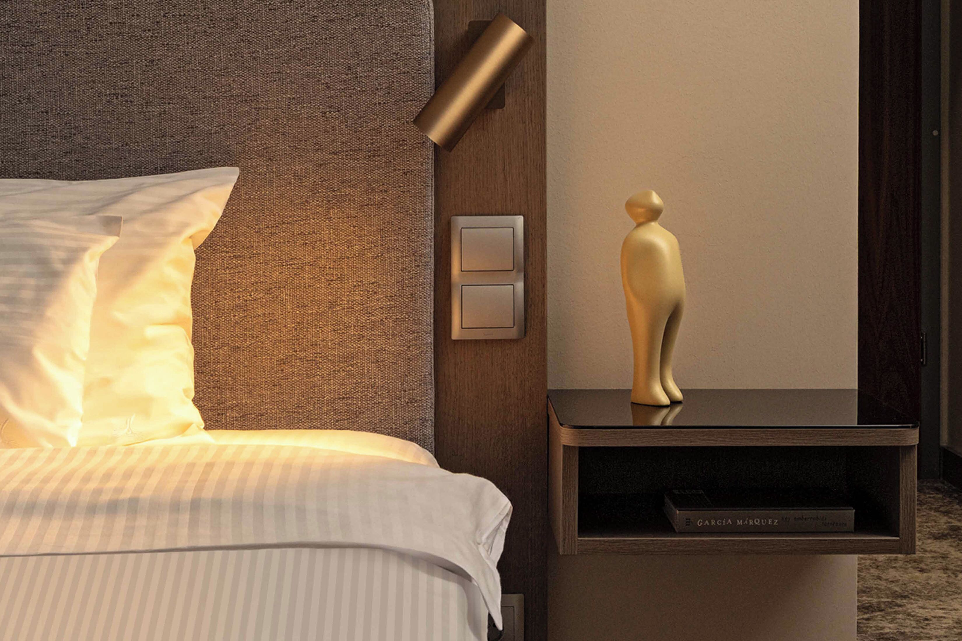 Una cama bien hecha con ropa de cama blanca, un cabecero texturizado, una lámpara de pared, una mesita de noche con un libro y una figura dorada decorativa.