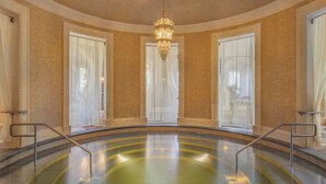 A large indoor swimming pool with a central chandelier, surrounded by walls with a textured finish and large windows with sheer curtains. - Spirit Hotel Thermal Spa (Sarvar)