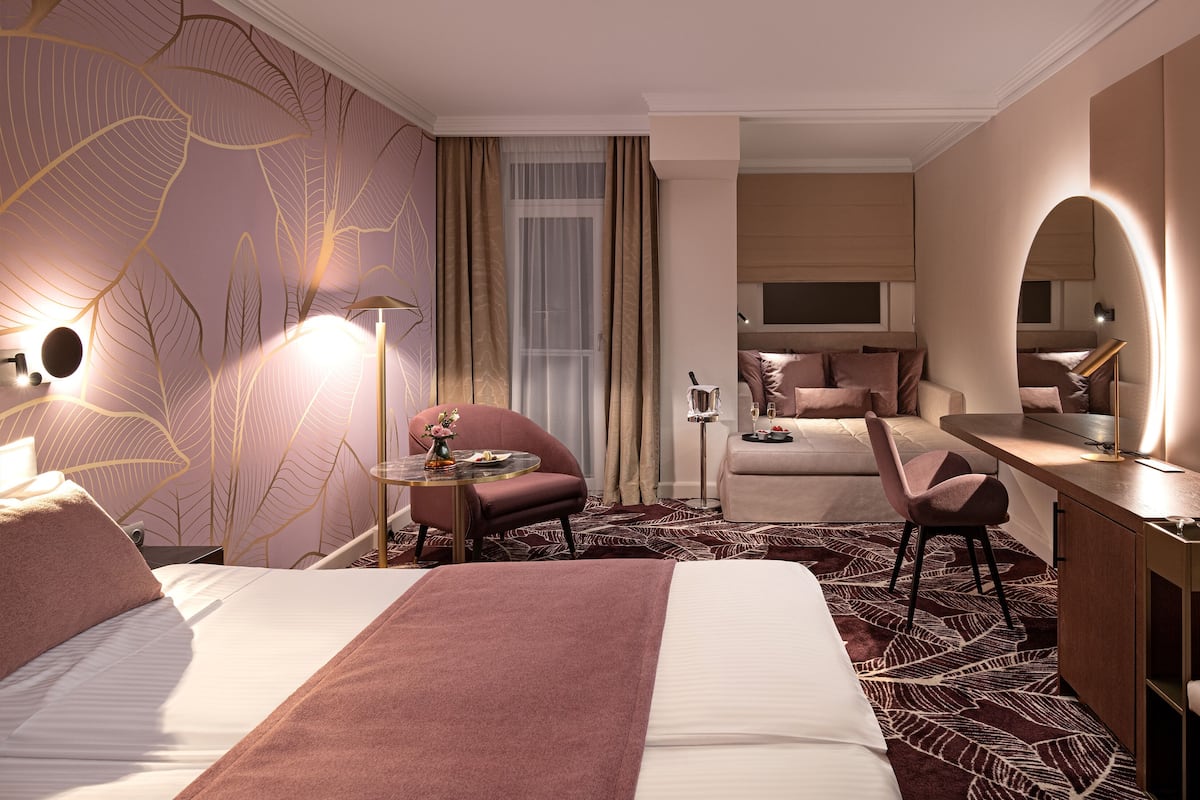 A hotel room with a large bed, a desk, a chair, a small table with a vase, and a mirror.