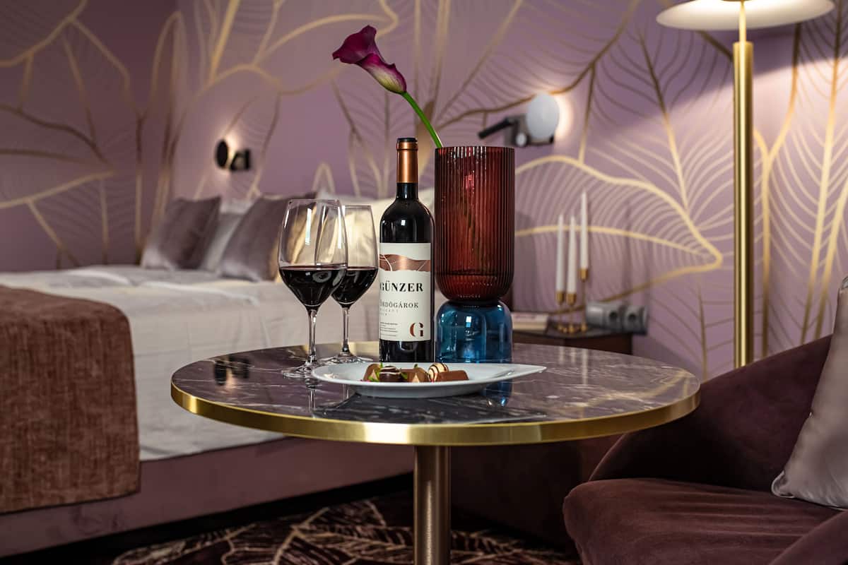 A hotel room with a bed, a round table with a bottle of wine, two glasses of wine, a plate with food, and a vase with a flower.