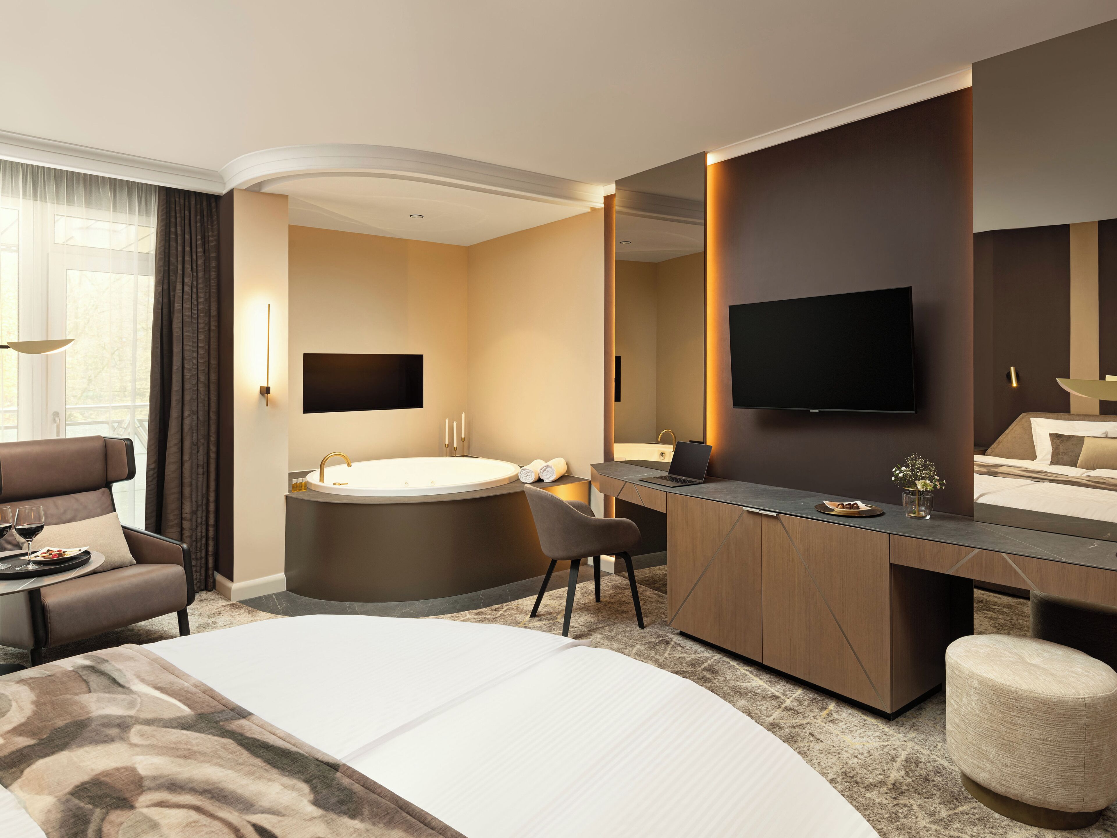 Una habitación de hotel moderna con una cama grande, un escritorio con una computadora portátil, una bañera y un televisor de pantalla plana montado en la pared.