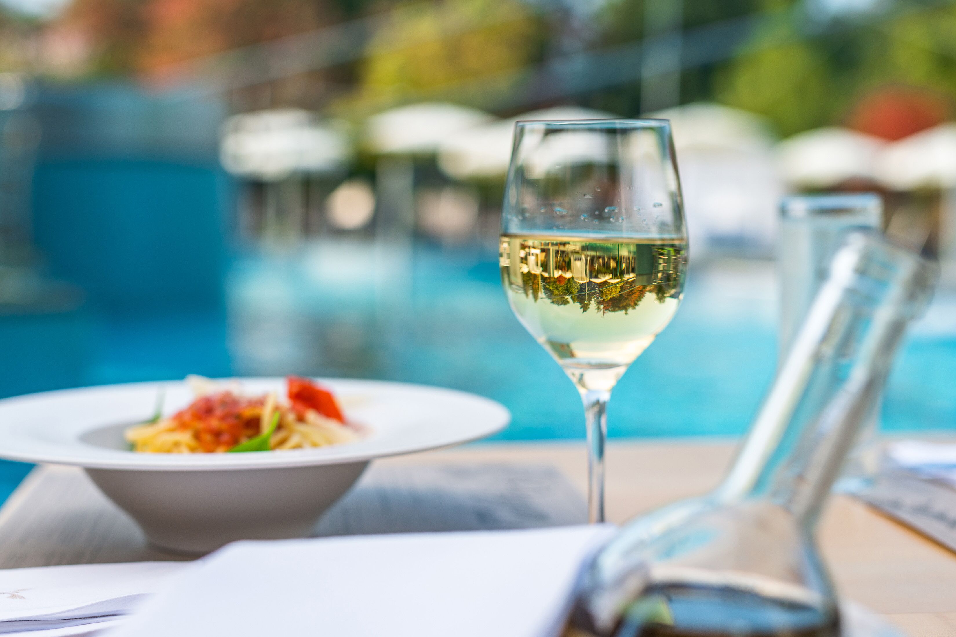 A dining table with a glass of white wine, a plate of food, and a pool in the background.