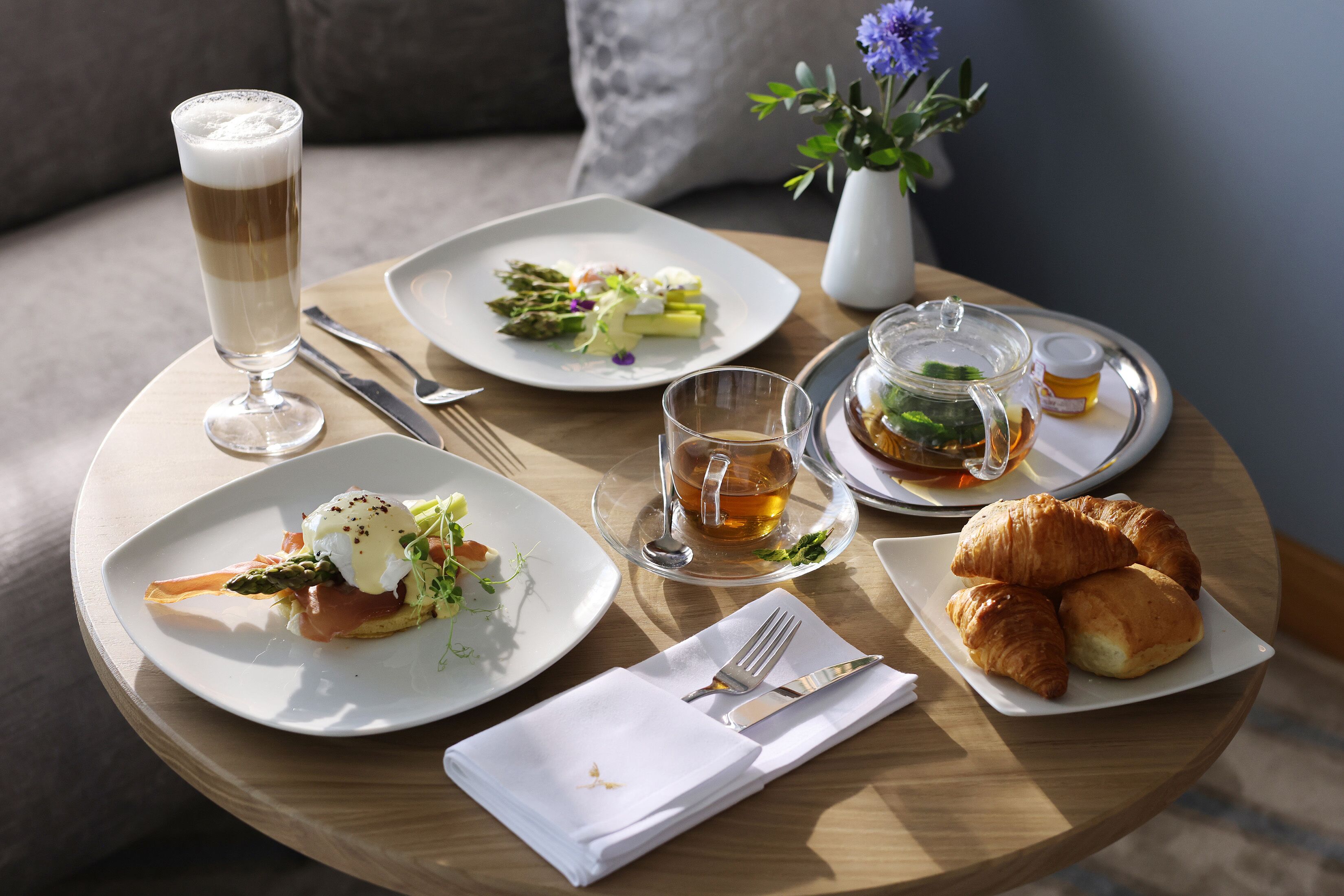 Desayuno con platos de comida, una taza de café, croissants y una tetera sobre una mesa redonda de madera.