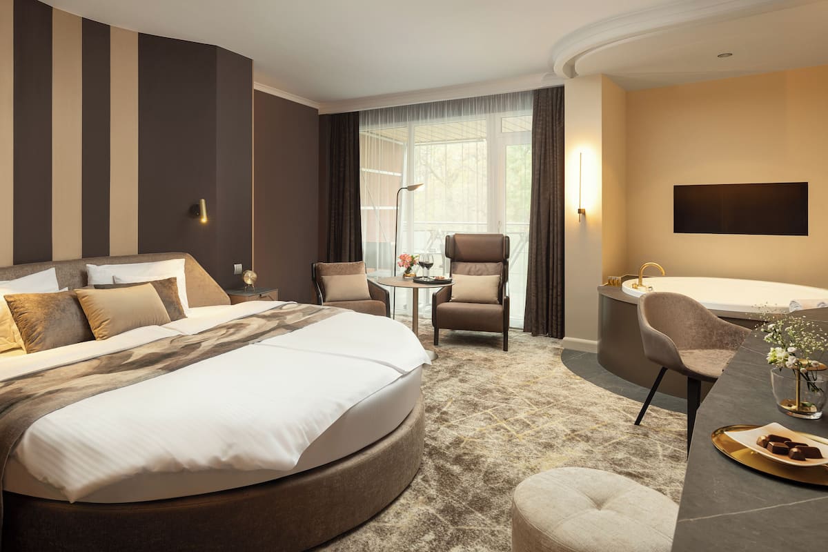 A hotel room with a large bed, a sitting area with a table and chairs, a desk, a bathtub, and a large window with curtains.