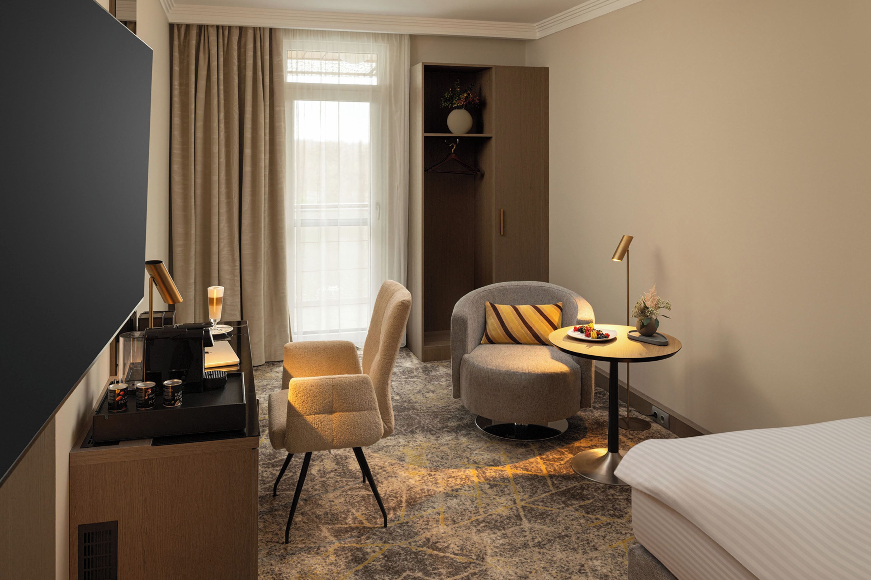A hotel room with a bed, a desk with a lamp, a chair, a small table with a lamp, and a wardrobe.