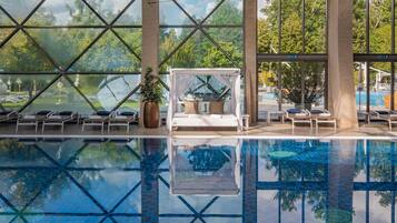 An indoor swimming pool with clear glass walls, lounge chairs, and a canopy bed.