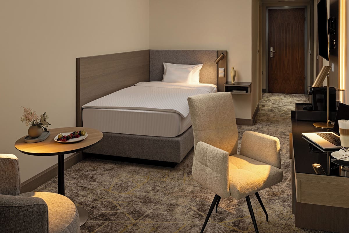 A hotel room with a bed, a desk, a chair, a small table with a vase and plate, and a television.