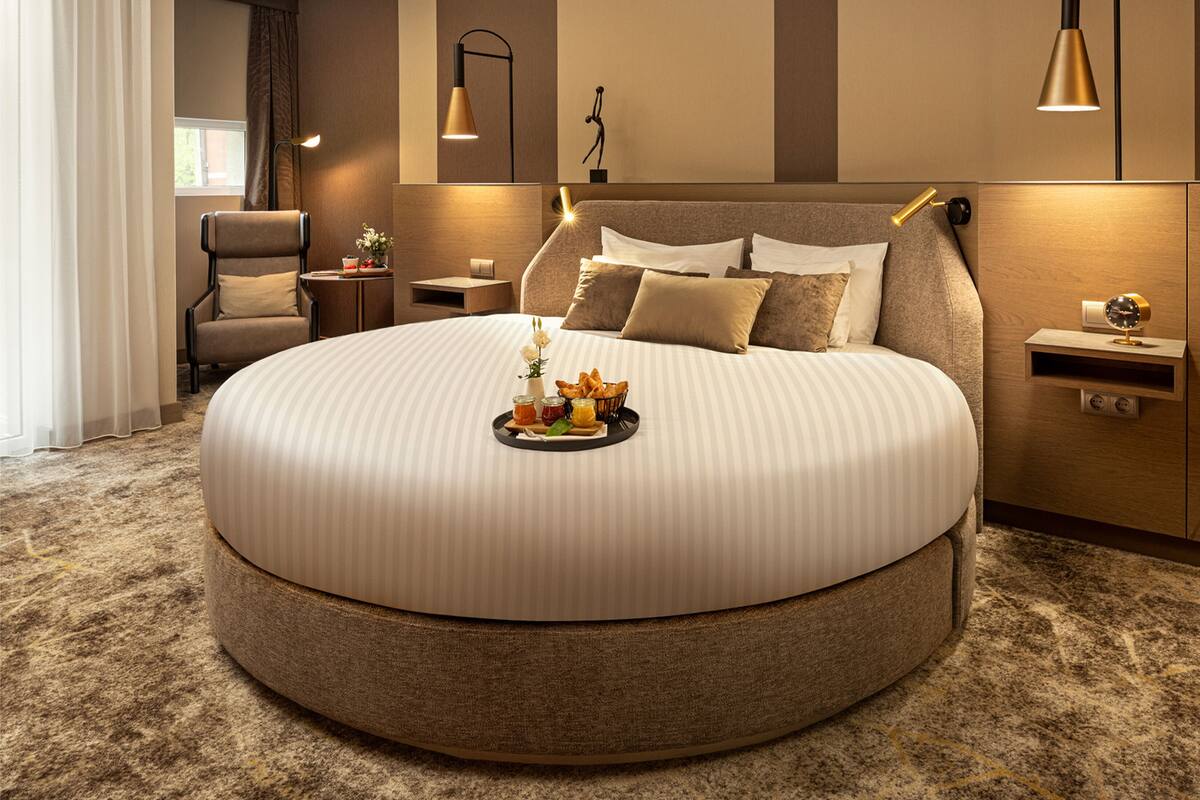 A round bed with a tray of food, a chair, bedside tables, and wall-mounted lamps in a modern hotel room.