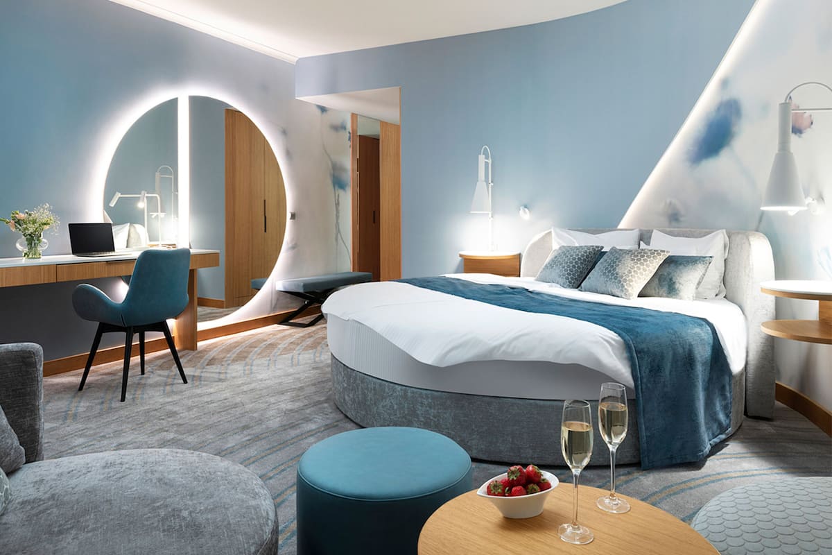 A modern hotel room with a large bed, a desk with a laptop, a round mirror, and a round table with a bowl of fruit and two glasses.