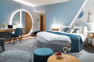 Una habitación de hotel moderna con una cama grande, un escritorio con una computadora portátil, un espejo redondo y una mesa redonda con un cuenco de fruta y dos vasos.