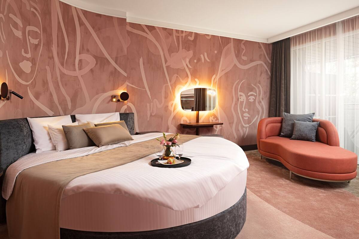 A modern hotel room with a large bed, a curved bedside table, a sofa, and a wall with abstract art.