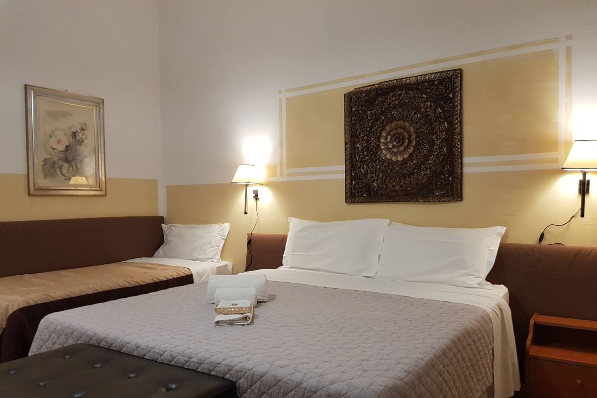A hotel room with two single beds, a large bed with a quilted bedspread, a bench, a wall-mounted artwork, and two wall lamps.