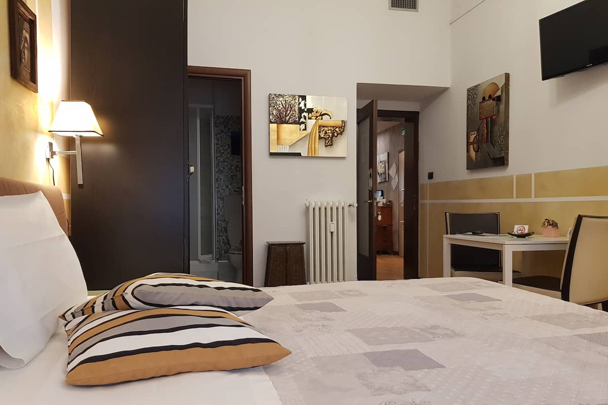 A hotel room with a bed, a desk, and a chair. There is a lamp on the wall and a painting on the wall.