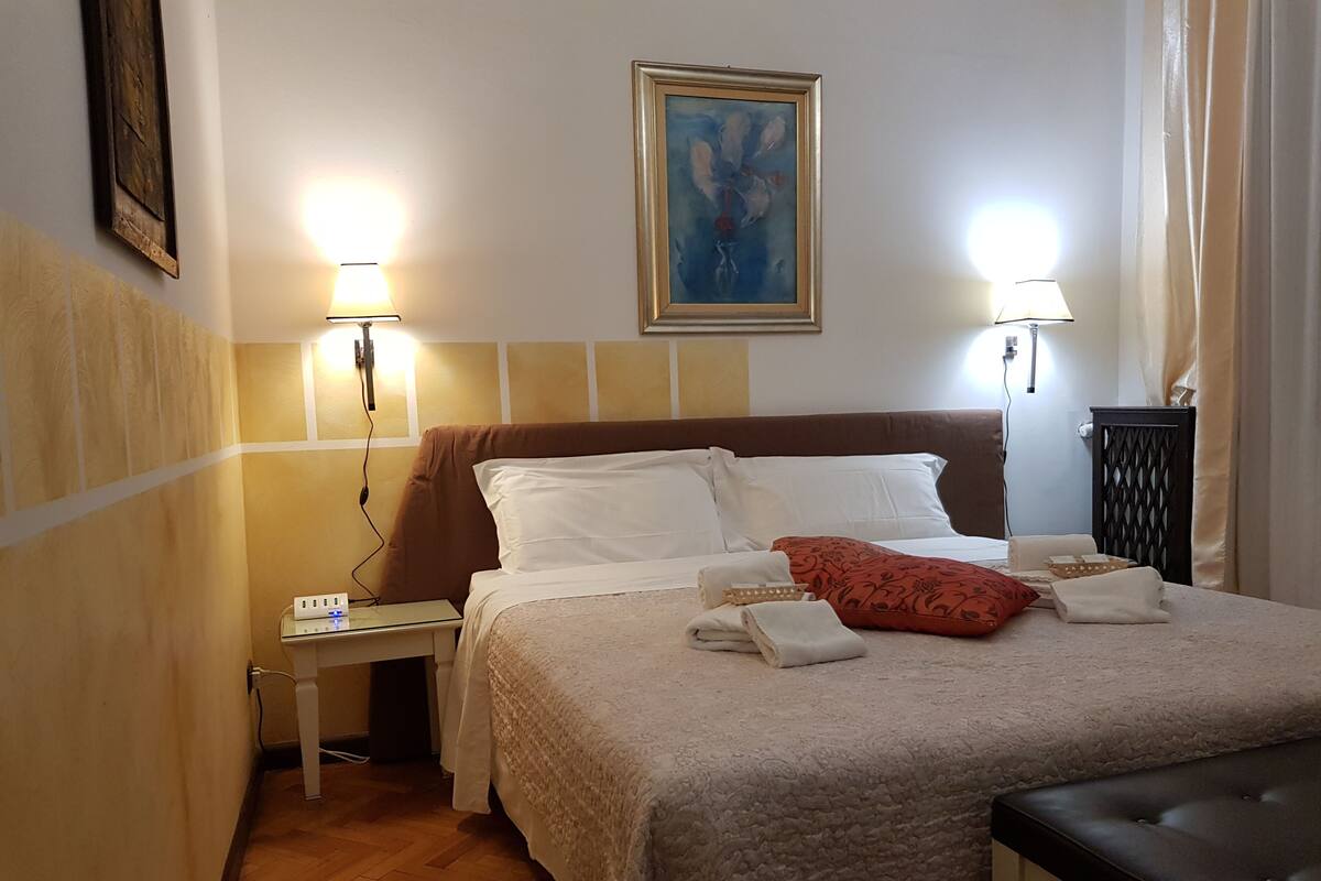 A neatly made bed with white linens, a brown headboard, and a red pillow. There are two bedside lamps, a small table with a lamp, and a painting on the wall.