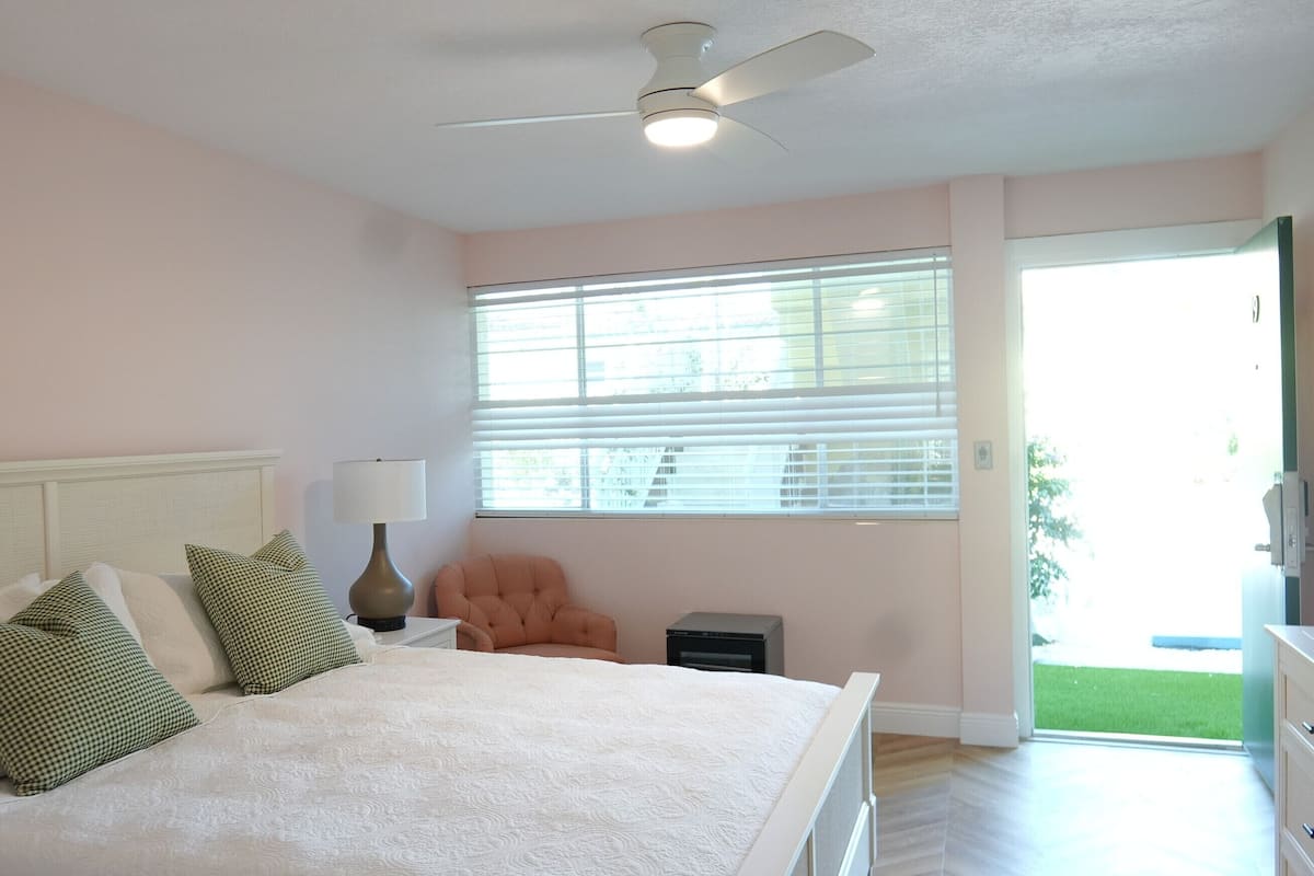 A bedroom with a bed, a chair, a lamp, a ceiling fan, and a window with blinds.