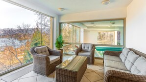 A balcony with wicker furniture, a glass table, and a view of a river and trees. - Triglav Bled (Bled)