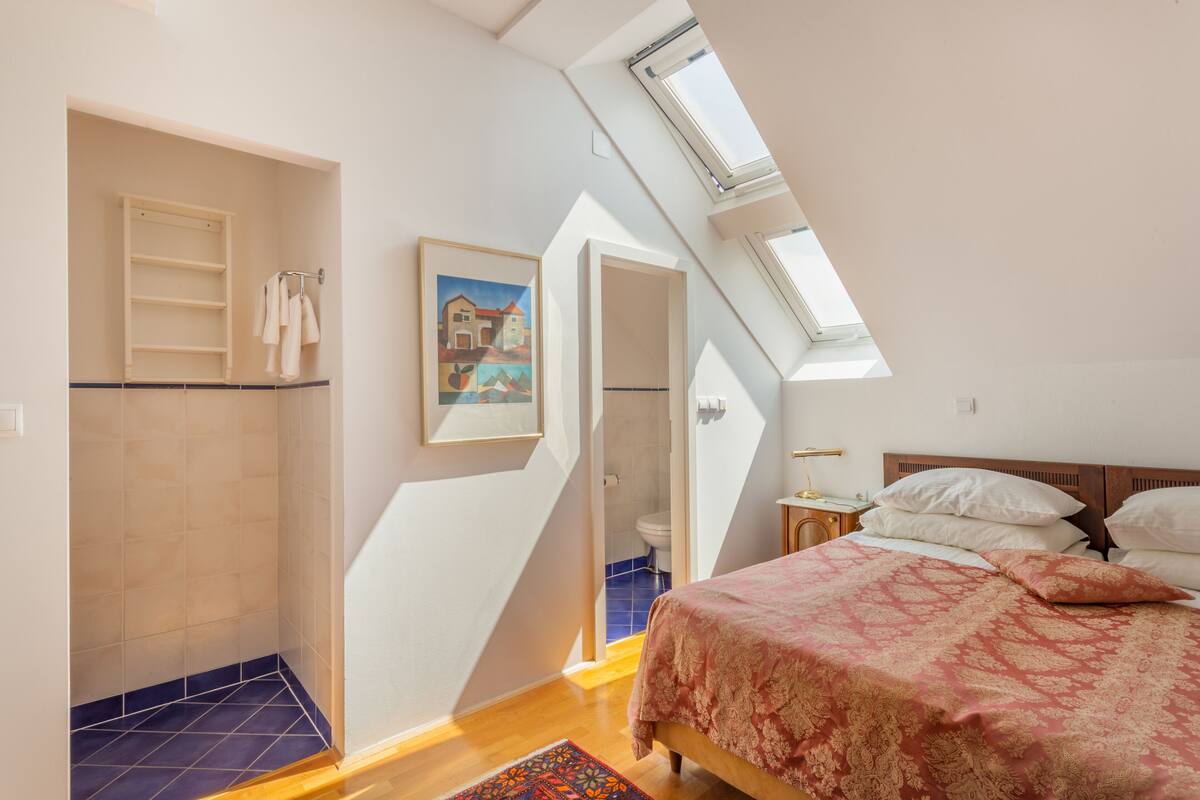 A bedroom with a skylight, a bed with a patterned bedspread, a painting on the wall, and a bathroom visible through an open door.