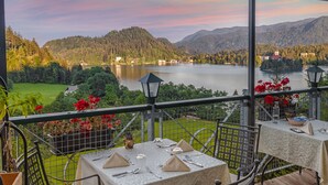 Un espacio al aire libre techado con zona de comedor y mesas dispuestas para dos, con vistas a un lago y colinas boscosas.