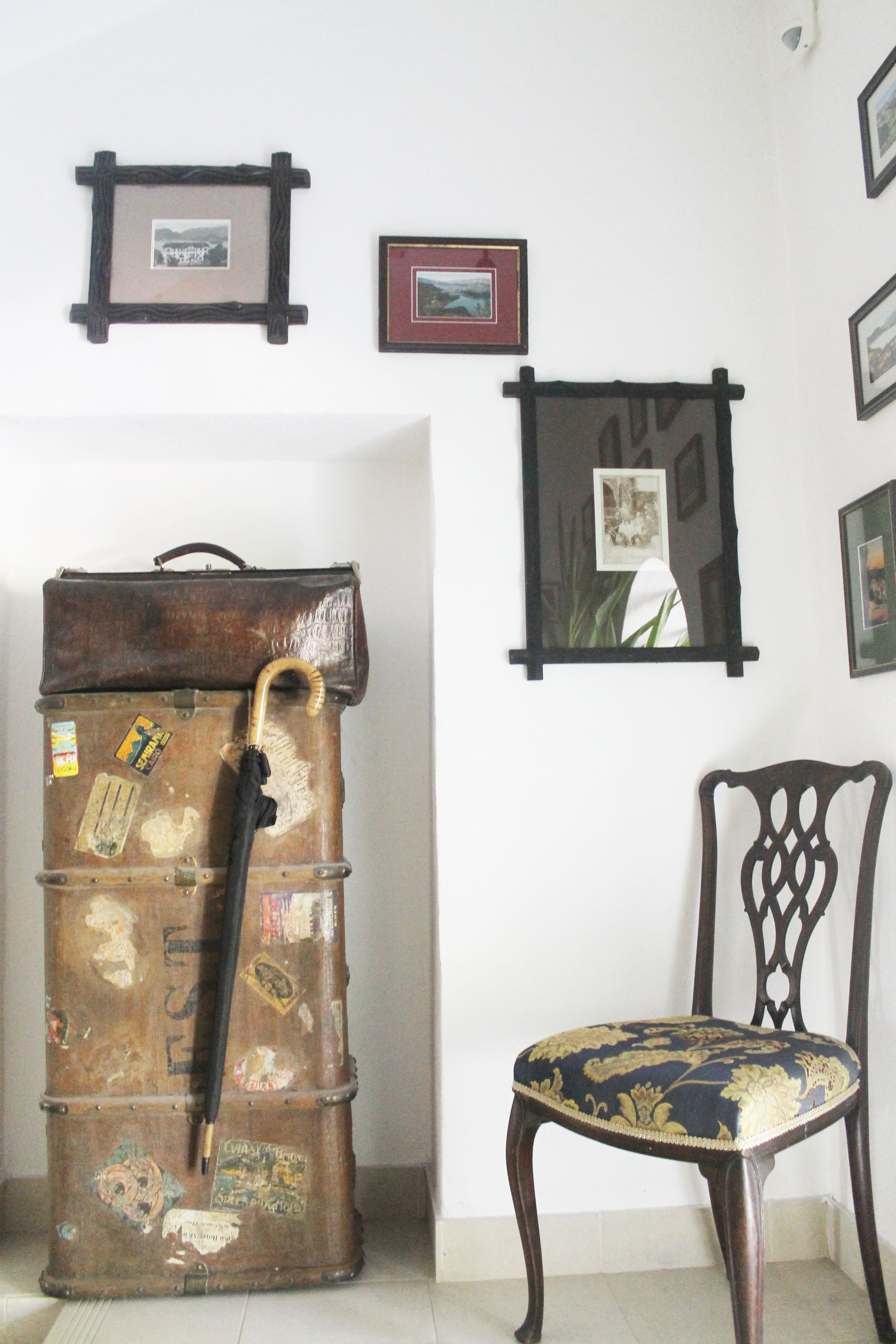 A vintage suitcase with a cane leaning against it, a wooden chair with a floral pattern, and framed pictures on the wall.