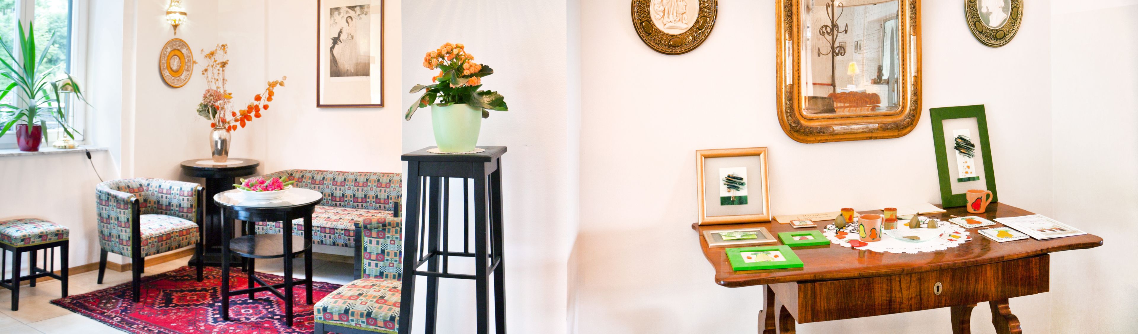 Una habitación con sillas estampadas, una pequeña mesa con un florero y un escritorio de madera con cuadros enmarcados y un espejo.