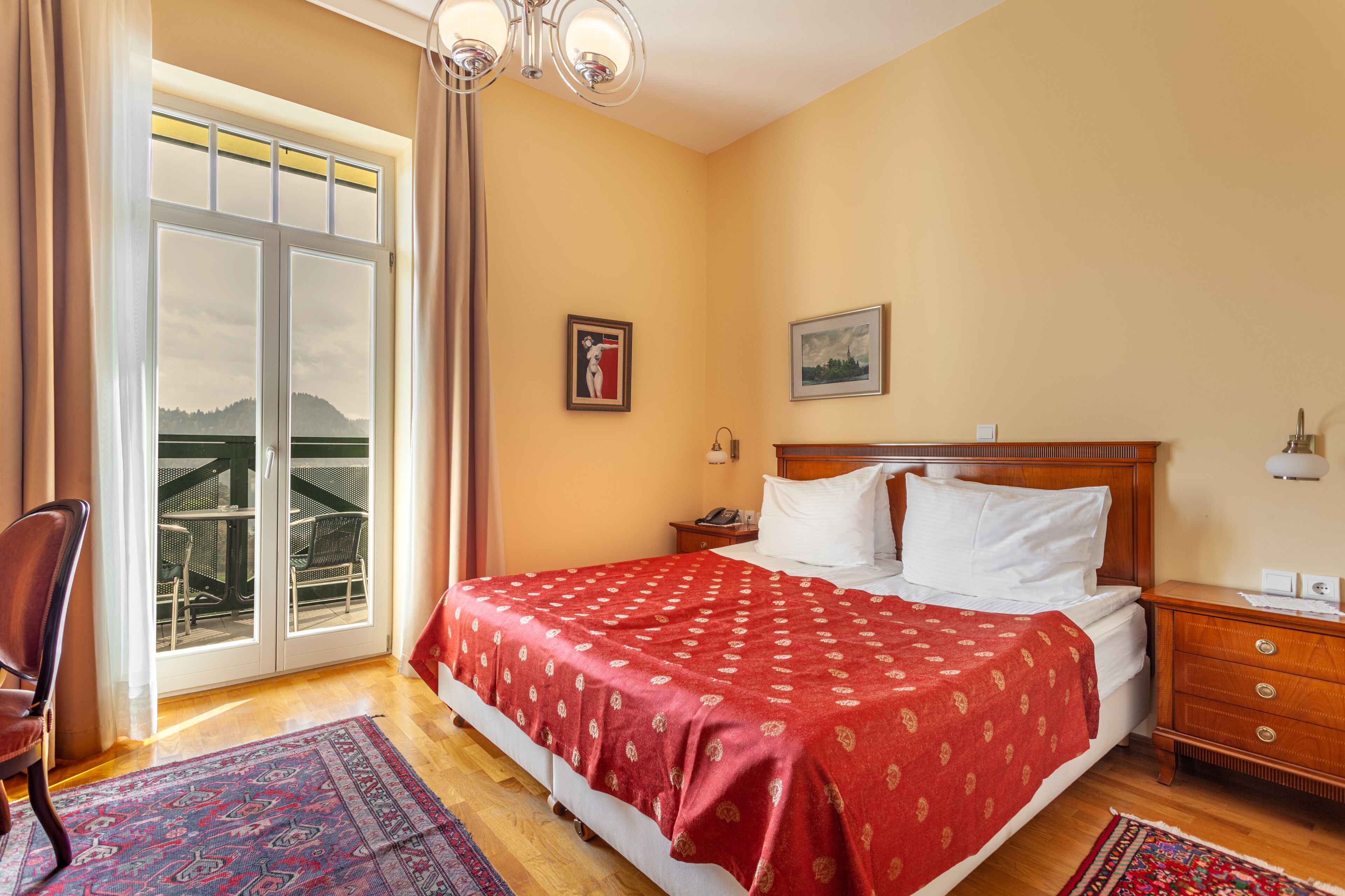 A bedroom with a large bed, a wooden nightstand, a desk, and a chair. There is a window with curtains, a painting on the wall, and a view of a bridge and hills outside.