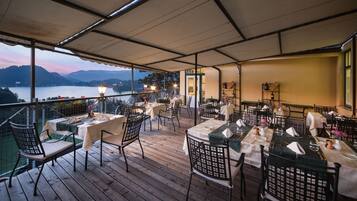 An outdoor dining area with tables and chairs, overlooking a body of water and hills.