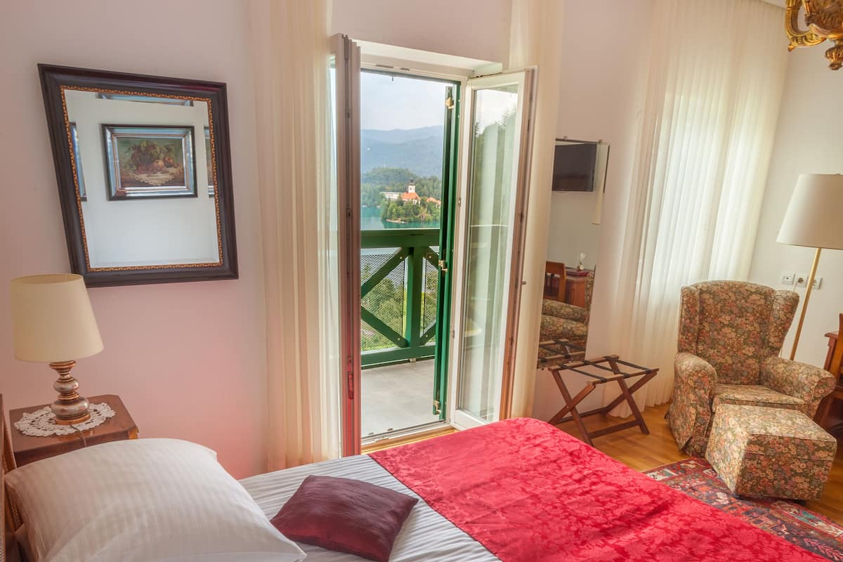 A hotel room with a bed, a chair, a table, and a view of a lake and mountains through an open door.