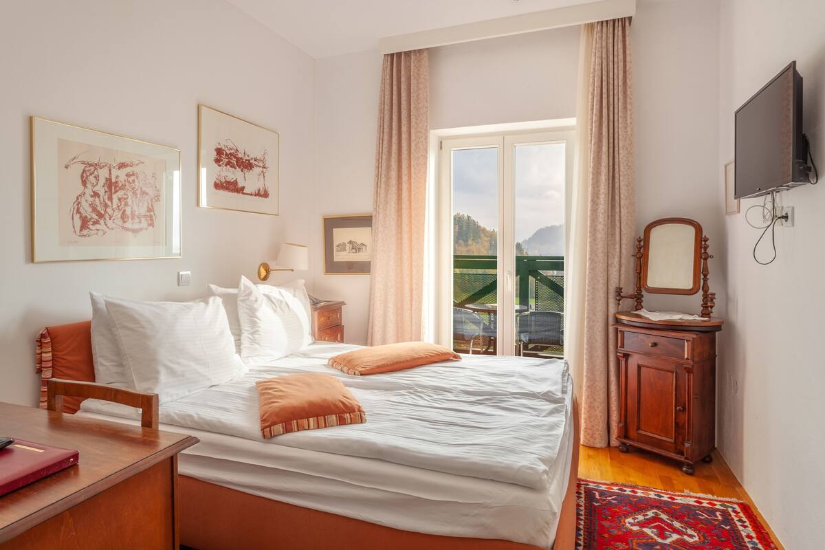 A bedroom with a bed, a wooden nightstand, a TV, and a window with a view of a mountain.