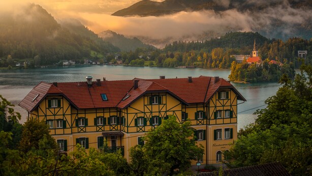 Un edificio tradicional con entramado de madera y techo rojo, ubicado junto a un lago con montañas al fondo.