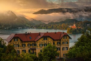 A traditional half-timbered building with a red roof, situated by a lake with mountains in the background. - Triglav Bled (Bled)