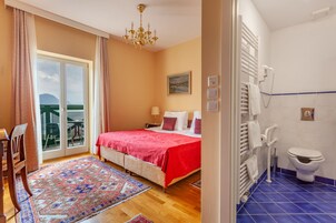 Un dormitorio con cama, mesita de noche, sillón, ventana con cortinas, araña y un cuadro en la pared, además de vistas al exterior.