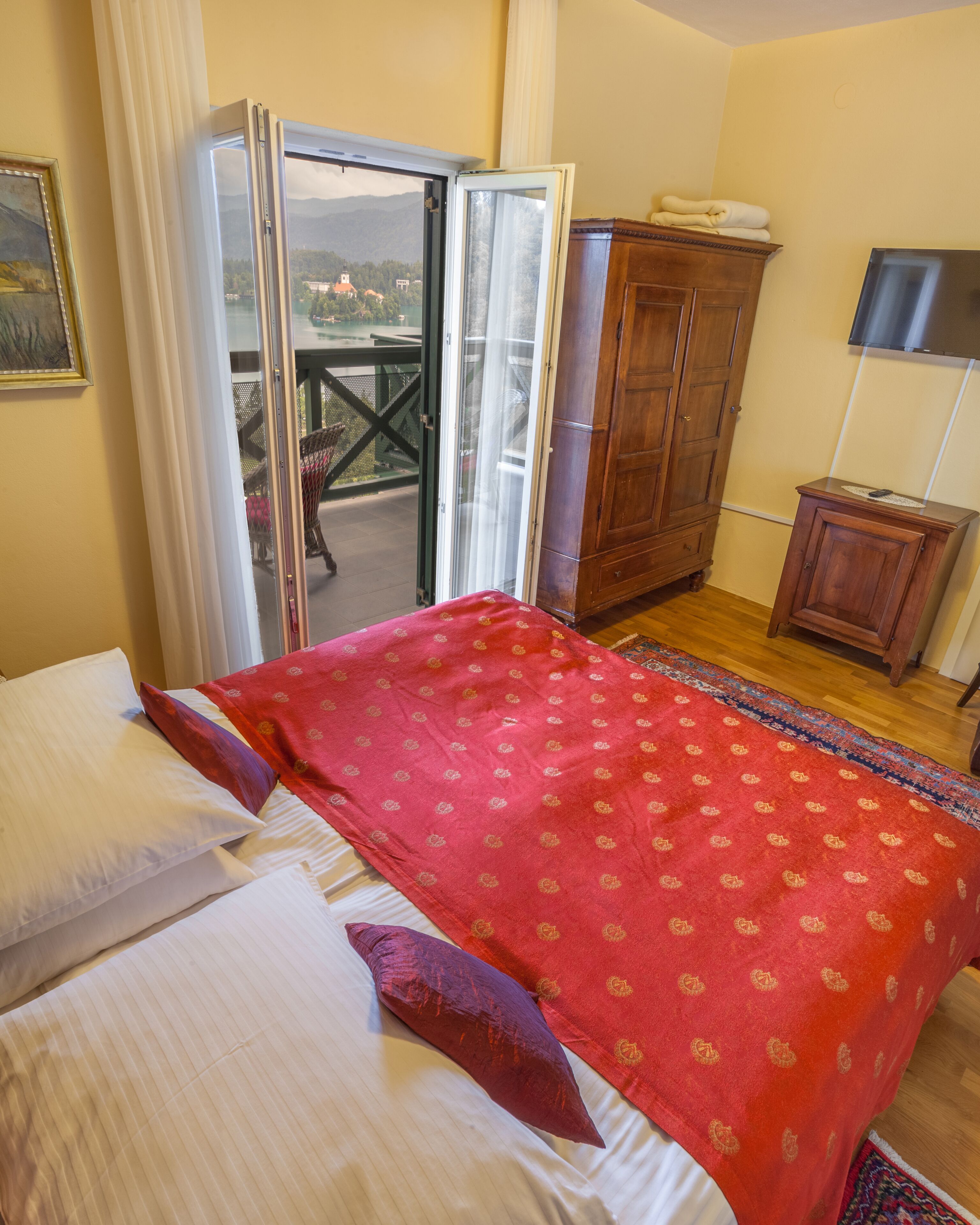Un dormitorio con cama, una colcha roja, un armario de madera, un mueble pequeño y un ventanal corredero que da a un área exterior.