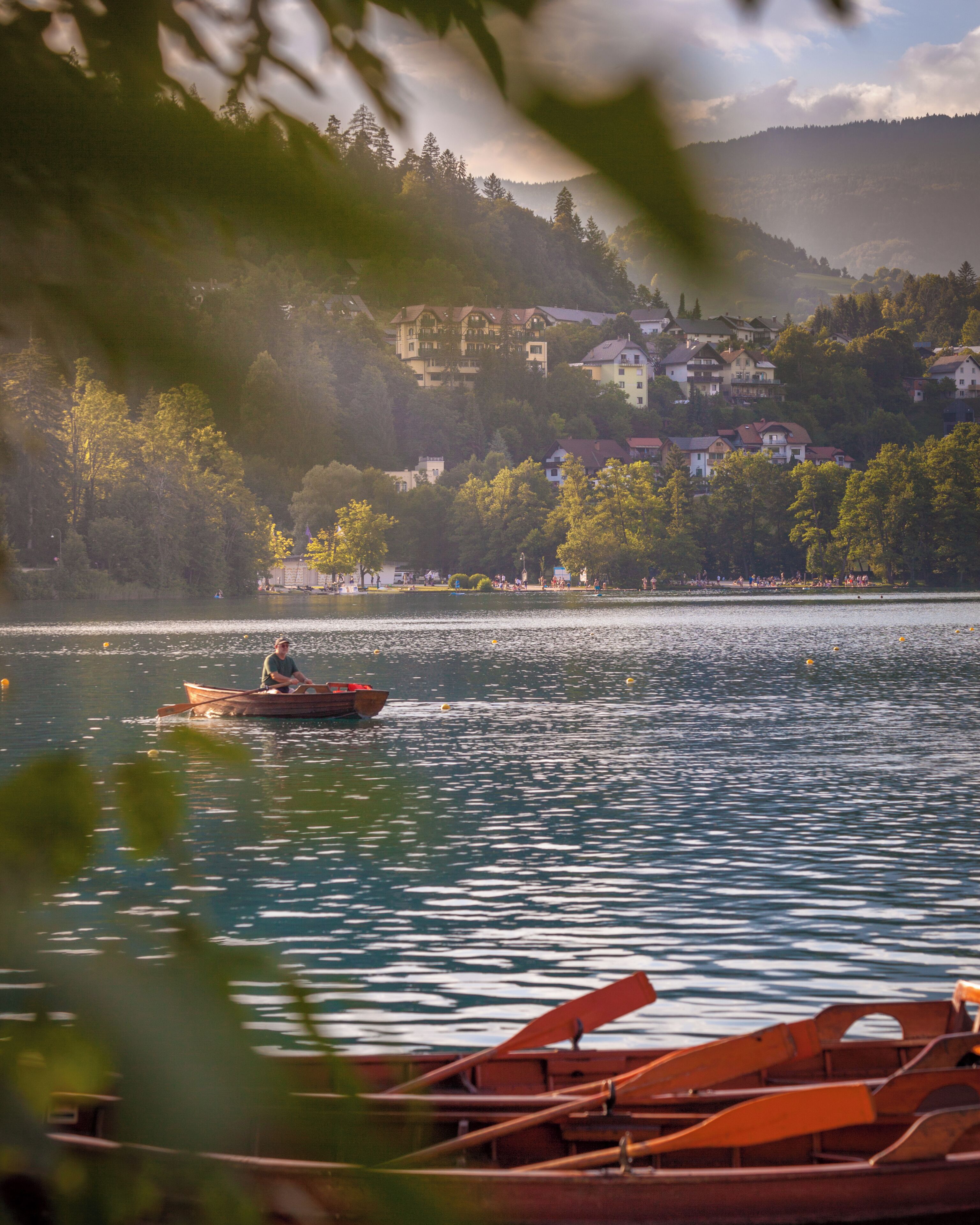 A calm lake with a rowboat in the foreground, surrounded by trees and houses in the background.