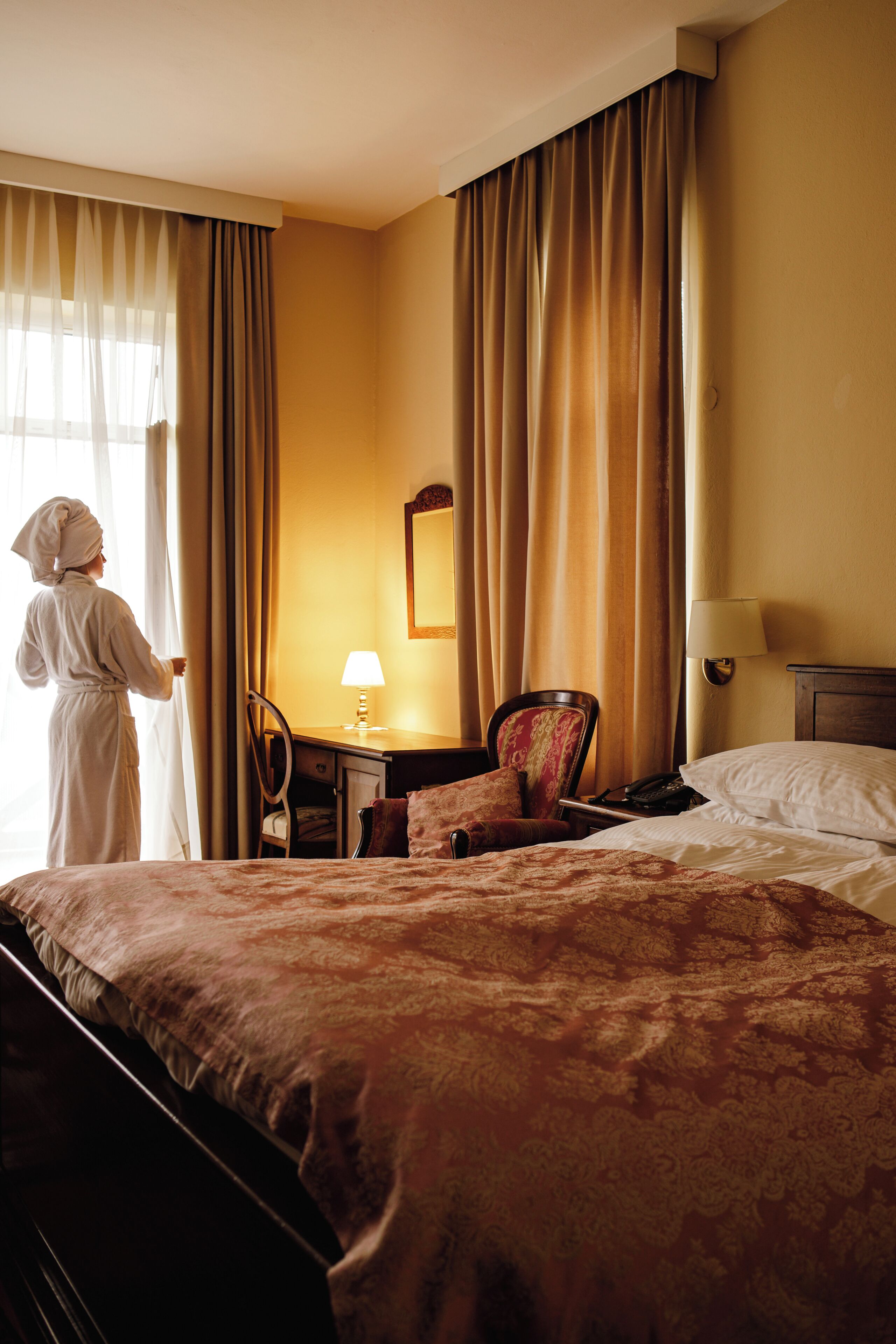 A hotel room with a bed, a chair, a desk, a lamp, and curtains.
