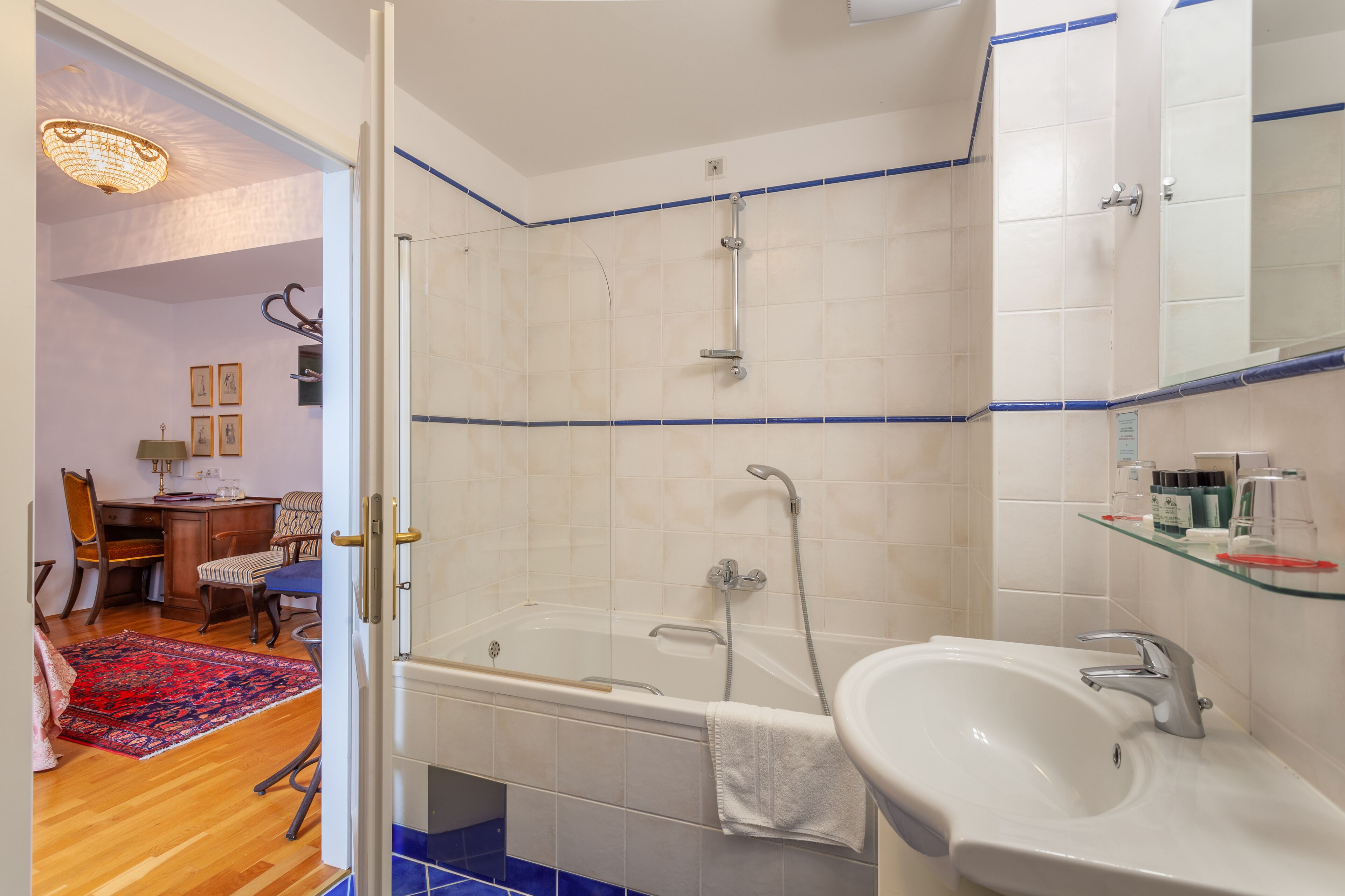 Un baño con bañera, lavamanos y ducha. Hay un estante de vidrio sobre el lavamanos y una toalla colgada en la pared.