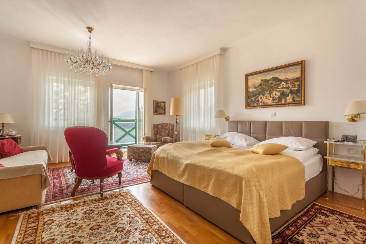 A hotel room with a large bed, a red armchair, a small table, and a painting on the wall.