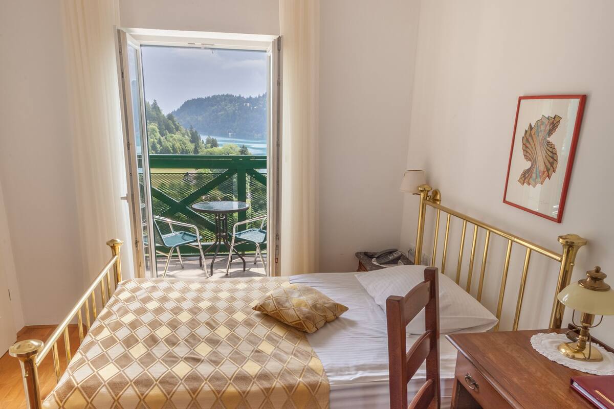 A bedroom with a bed, a desk, a chair, a window with a view of greenery, and a chandelier.