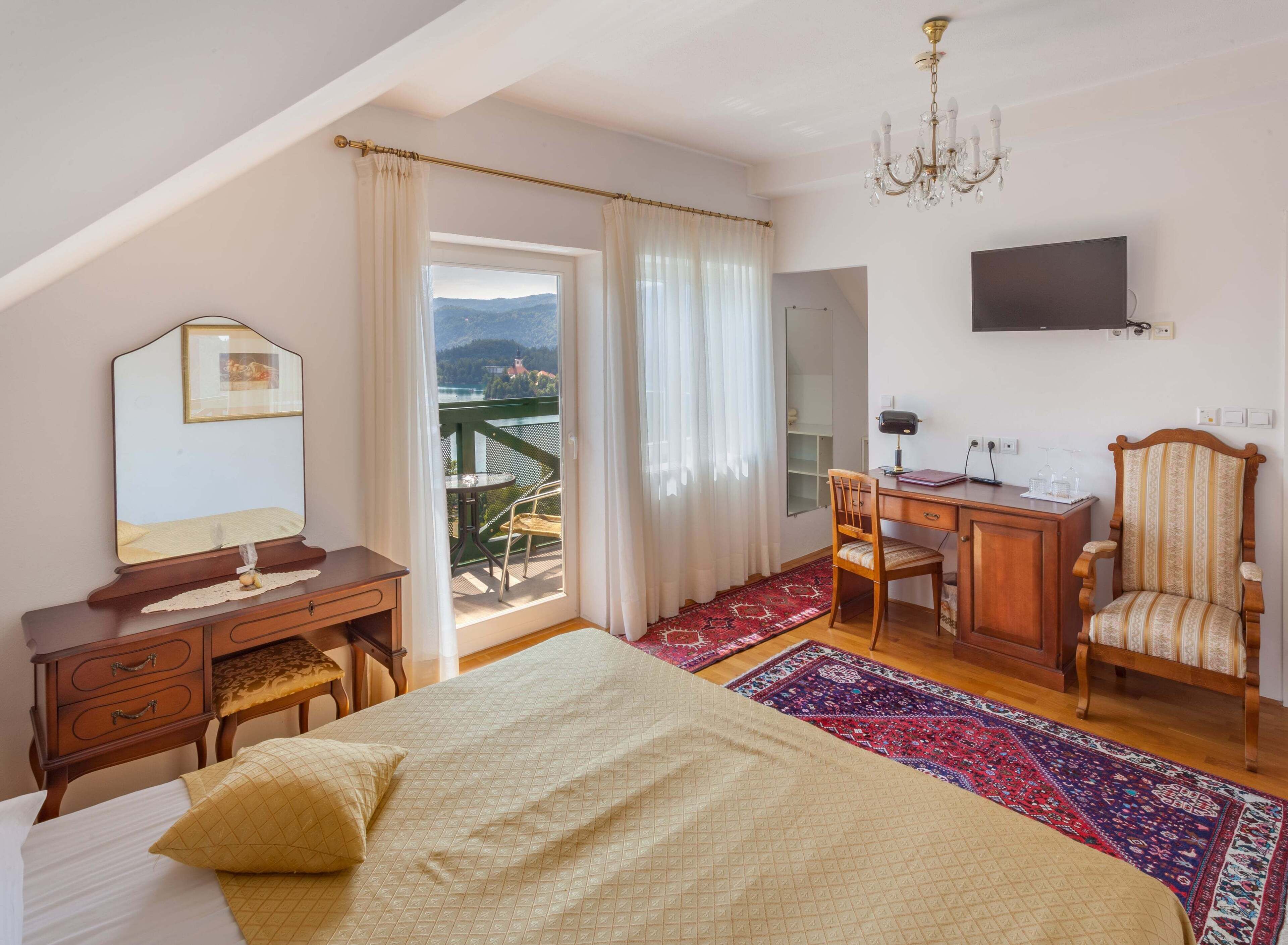 A bedroom with a bed, a desk, a chair, a TV, and a balcony with a view.