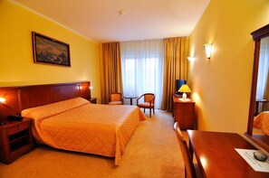 A hotel room with a large bed, two chairs, a small table, a desk, a lamp, a painting on the wall, and a window with curtains. - Maxim Oradea (Oradea)