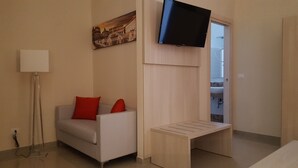 Habitación de hotel moderna con sofá, una pequeña mesa, televisor de pantalla plana montado en la pared y un cuadro de un edificio histórico.