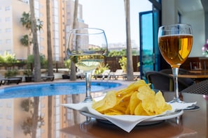 A table with a plate of chips and two glasses of wine, a pool, and a view of a building with palm trees. - Hotel Cibeles Playa (Gandia)