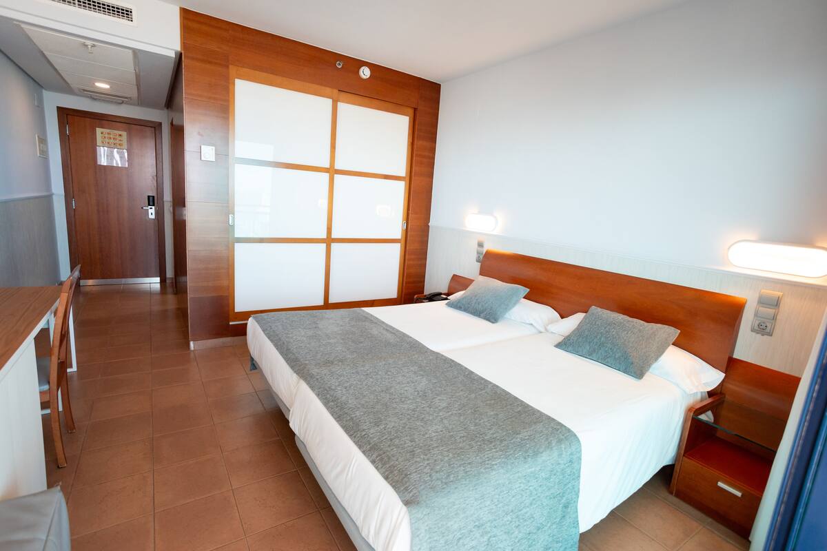 Quarto de hotel com cama grande, cabeceira de madeira, mesa de cabeceira com abajur, guarda-roupa e janela com persianas.