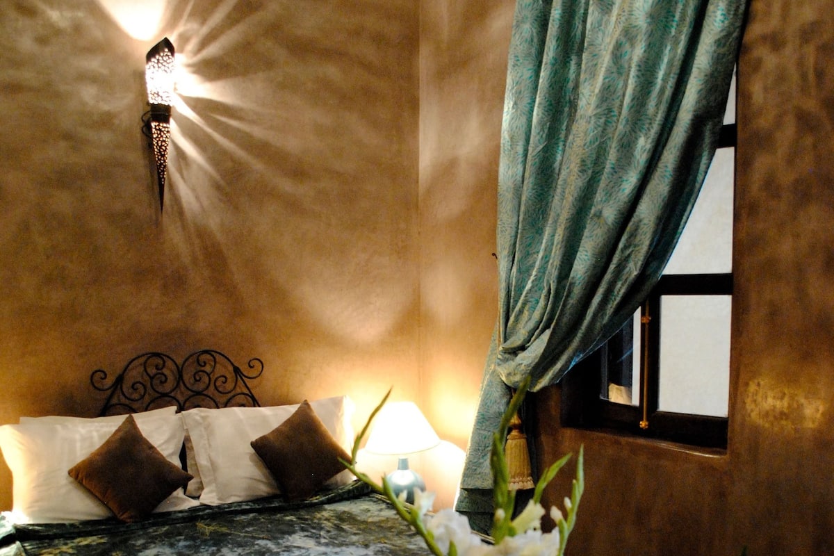 Un dormitorio con cama, almohadas, un florero con flores, una lámpara y una ventana con cortinas.