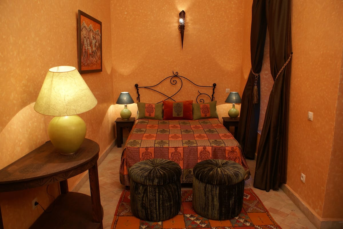 Un dormitorio con una cama, dos mesitas de noche, una lámpara, una luz empotrada en la pared y una alfombra estampada.