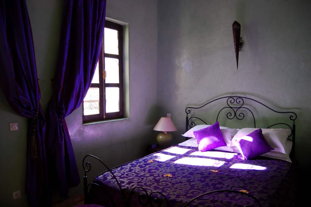 Un dormitorio con cama, cortinas moradas, una ventana y una lámpara fijada a la pared.